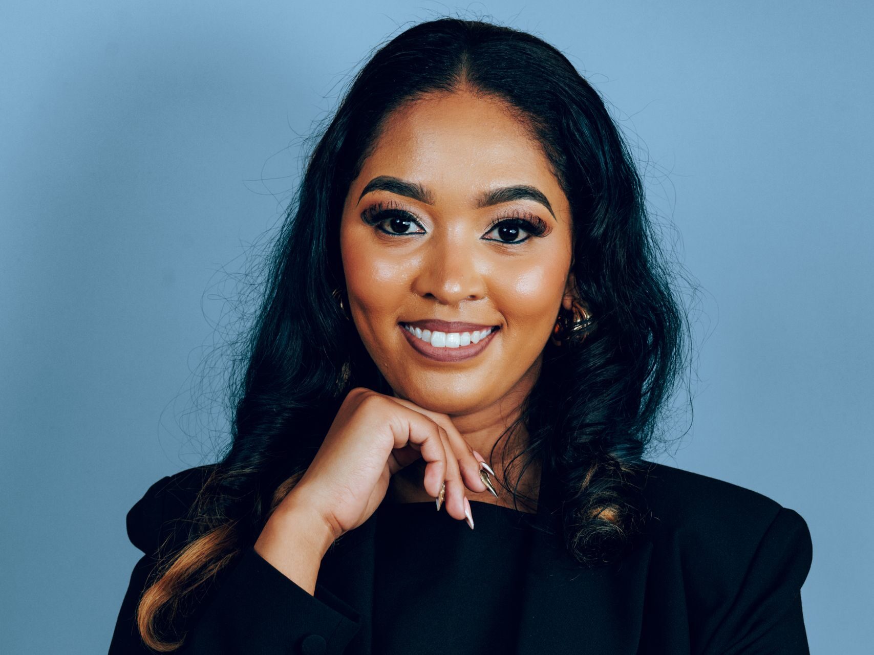 Confident woman in a black suit poses against a blue background, exuding professionalism and charm.