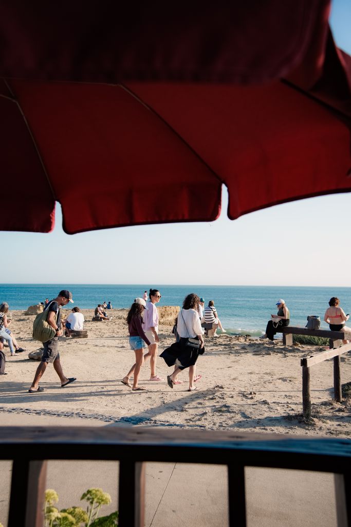 People walking along a sunny beach, captured under a red umbrella, with a calm ocean view.