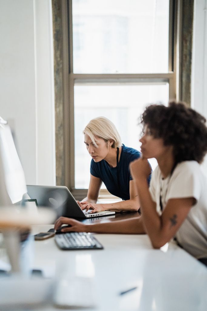 Two professional women collaborating at an office desk using laptops, focused on work.